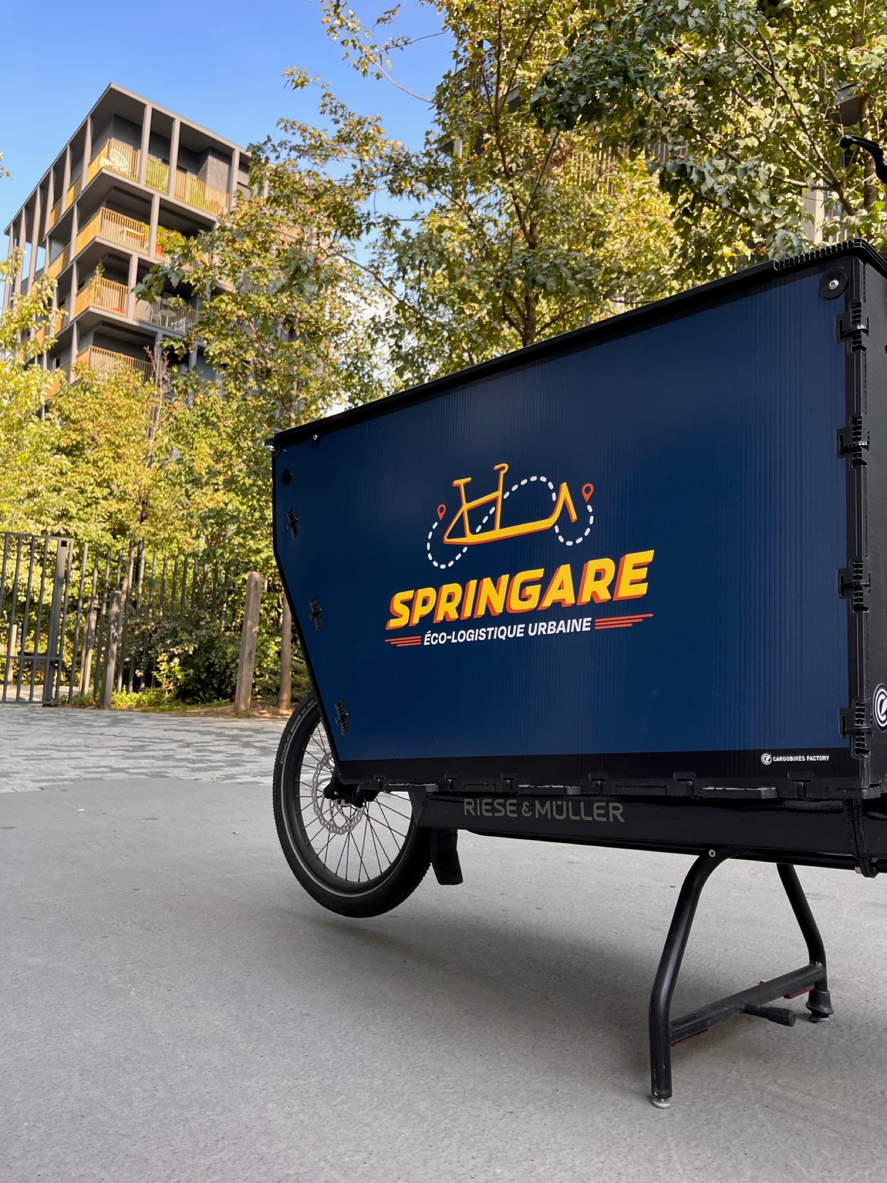 La Cargo Box Springare bleue, montée sur un vélo cargo Riese & Müller, dans une rue plantée d'arbres