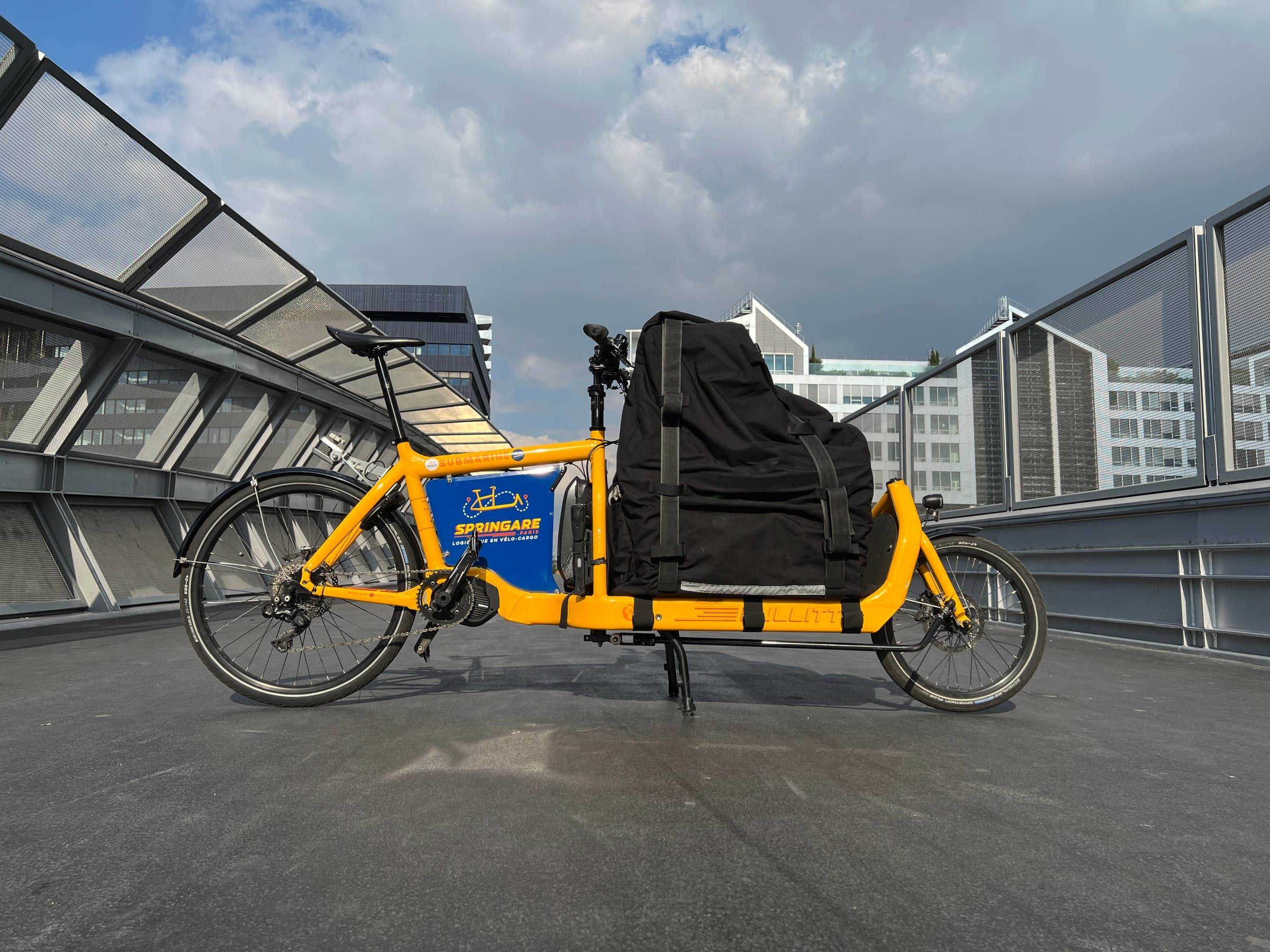 Le Cargo L, vélo cargo jaune Springare chargé d'un grand sac étanche noir, sur une passerelle