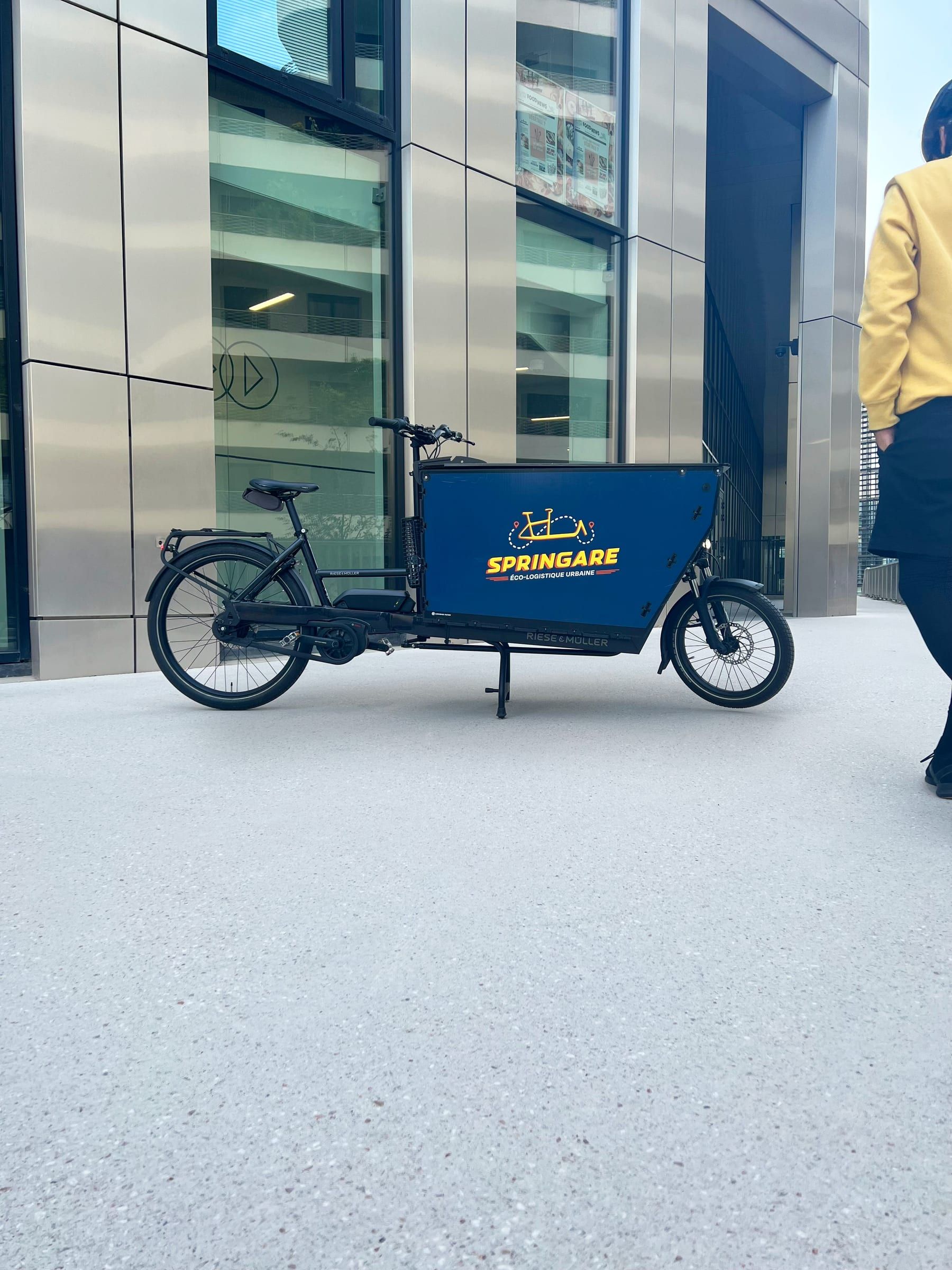 Le Cargo XL, vélo cargo Riese & Müller à caisson bleu Springare, garé devant un immeuble de bureaux