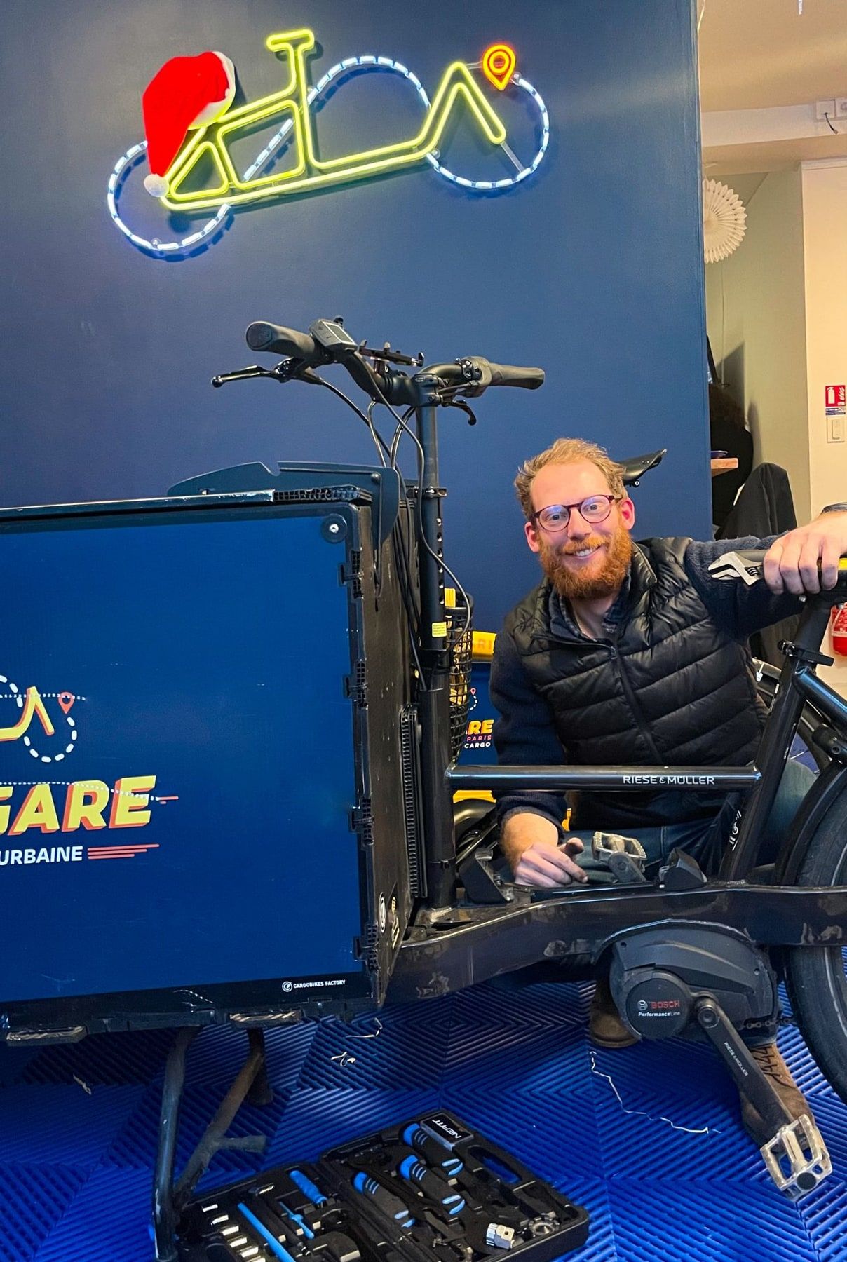 Geoffroy, mécanicien, accroupi derrière un vélo cargo démonté, sous l'enseigne néon en forme de vélo de l'atelier