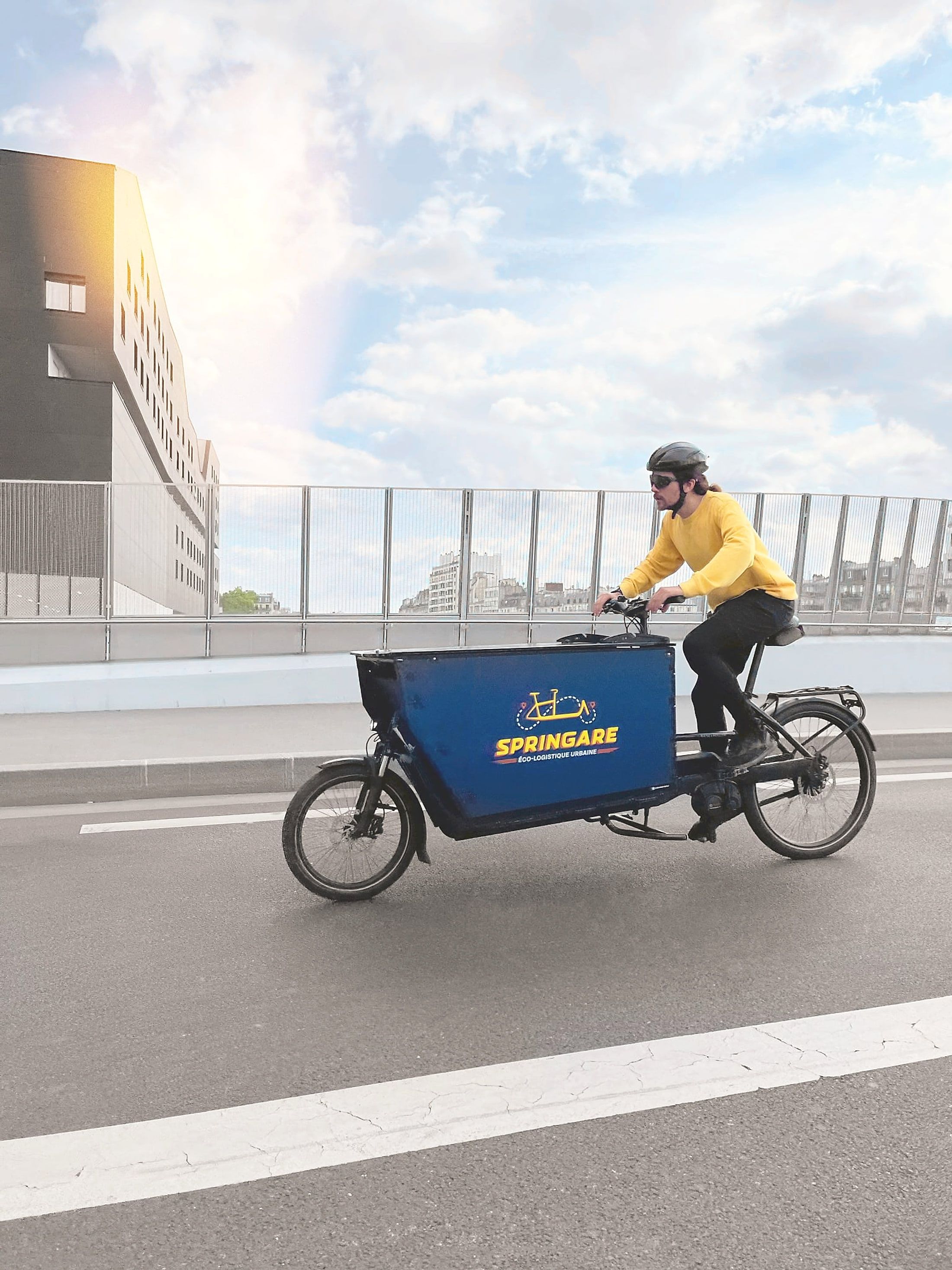 Un coursier Springare en pull jaune roule à vélo cargo sur un pont parisien