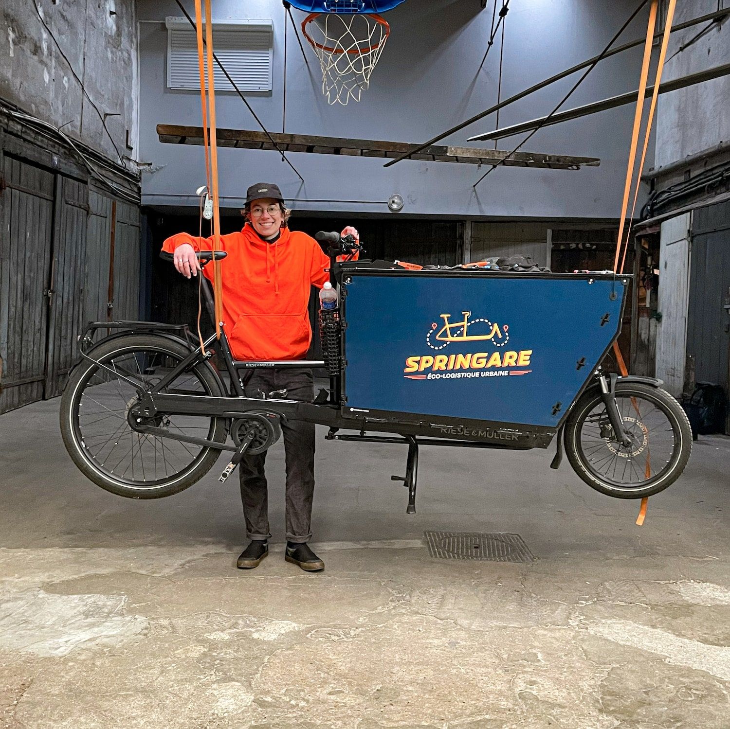 Juliette, en sweat orange, appuyée sur le guidon d'un vélo cargo dans l'atelier
