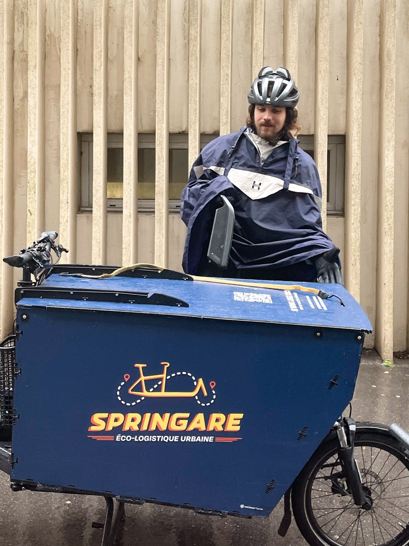 Un coursier en poncho de pluie et casque, derrière le caisson bleu de son vélo cargo