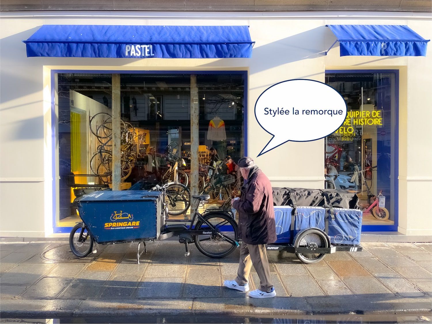 Un vélo cargo attelé à sa remorque devant la boutique Pastel, avec une bulle « Stylée la remorque »