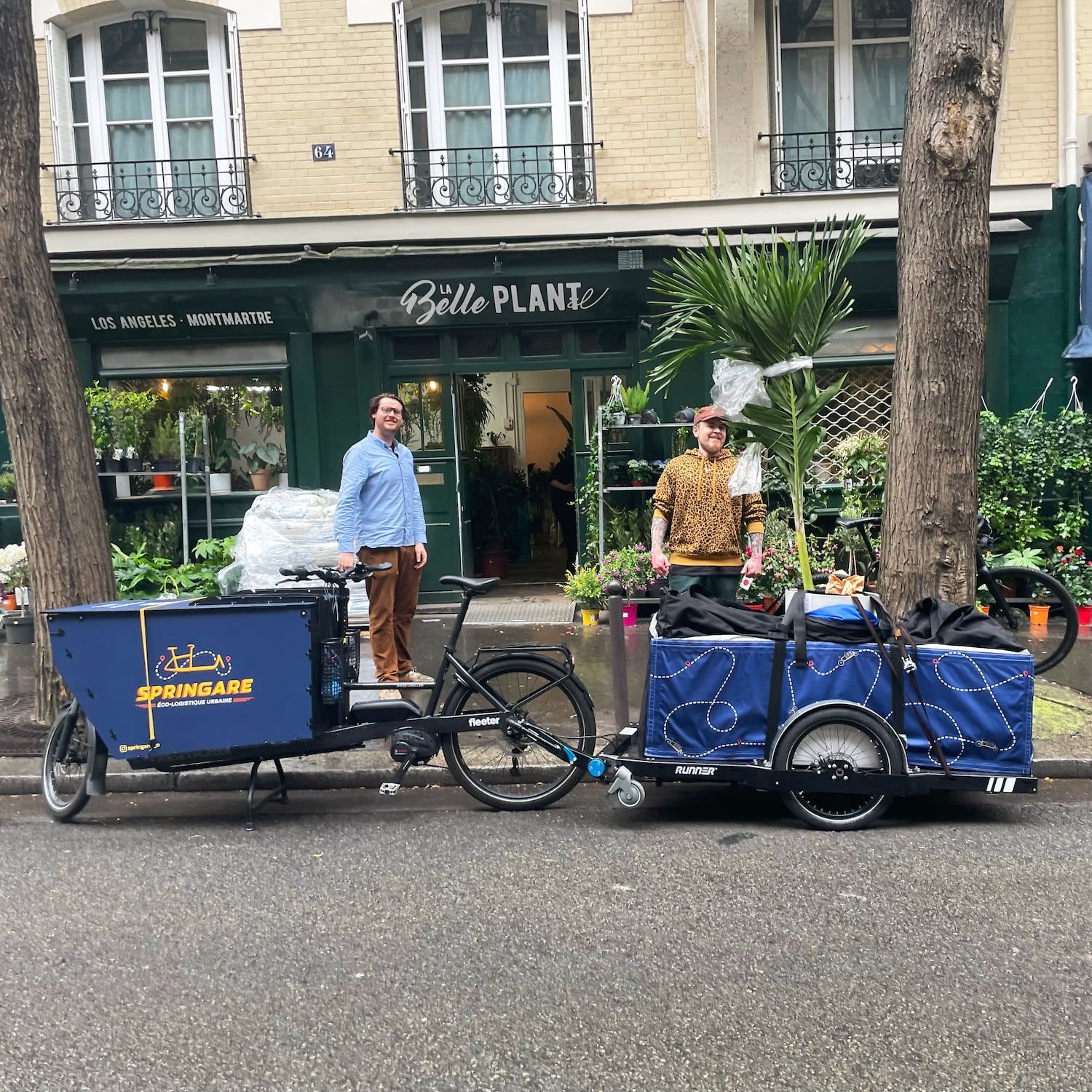Un palmier calé dans la remorque Springare devant la boutique La Belle Plante, deux hommes posant de part et d'autre