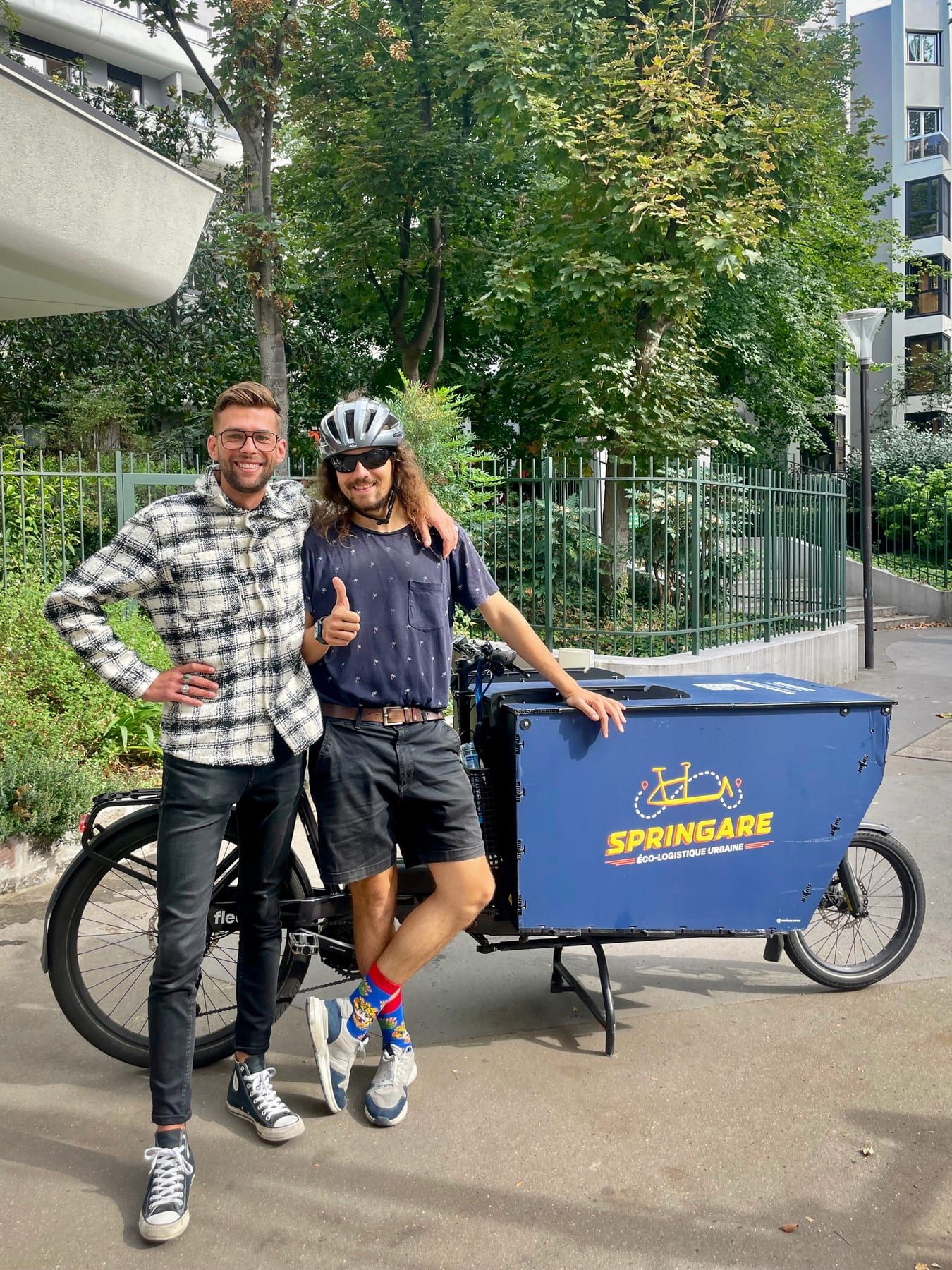 Deux hommes posent en souriant, pouce levé, à côté d'un vélo cargo Springare dans une rue arborée